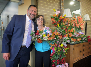 Welcome to Easter Services - Rev Russ and Kate Law smile their welcomes next to the beautiful flowers on the Easter Cross